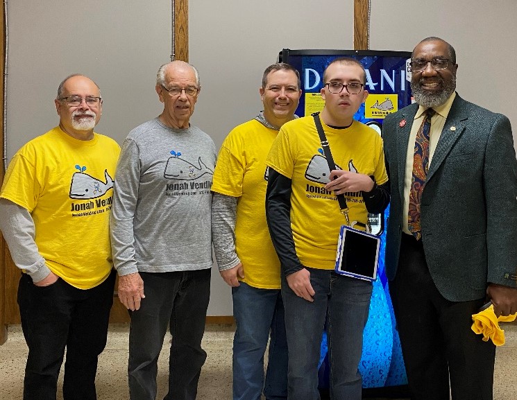 Three men wearing yellow shirts with "Jonah Vending" logo standing with self-advocate Jonah Taylor and State Rep. Jerome Barnes. 